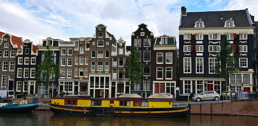 A row of brick rowhouses  along a canal: most are quite narrow with a decorative gable and many large windows. A bright yellow houseboat moored in the canal.
