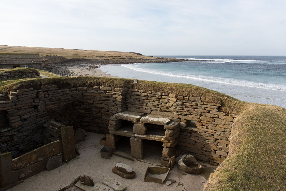 Stone walls in curved shape show the insides of a room, turf shows at the top of the walls. Something like an oven made of stones against the wall inside. Beyond the walls, a beach with a calm sea.
