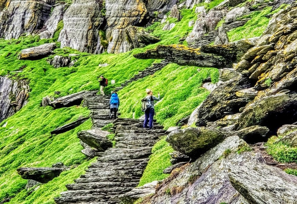 On a steep, rocky hill, a rough stone stairway. A few people climb it.