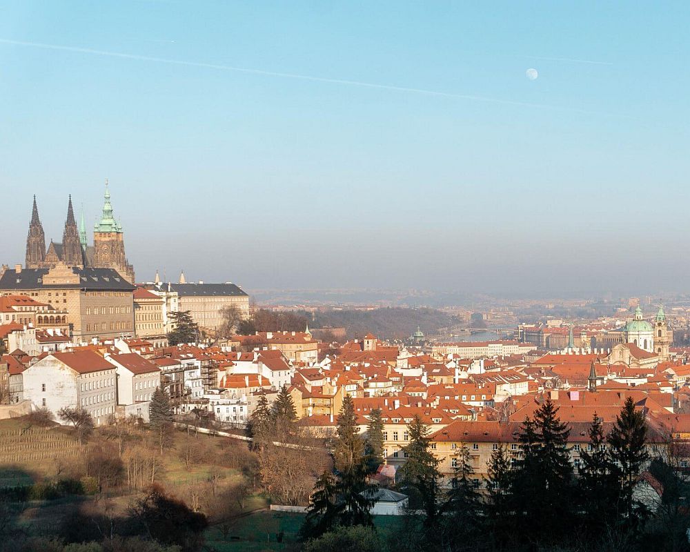 A view from a hill looking down over the city. To the left, the spires of Prague Castle. Ahead, the red-tiled roofs of the city. 