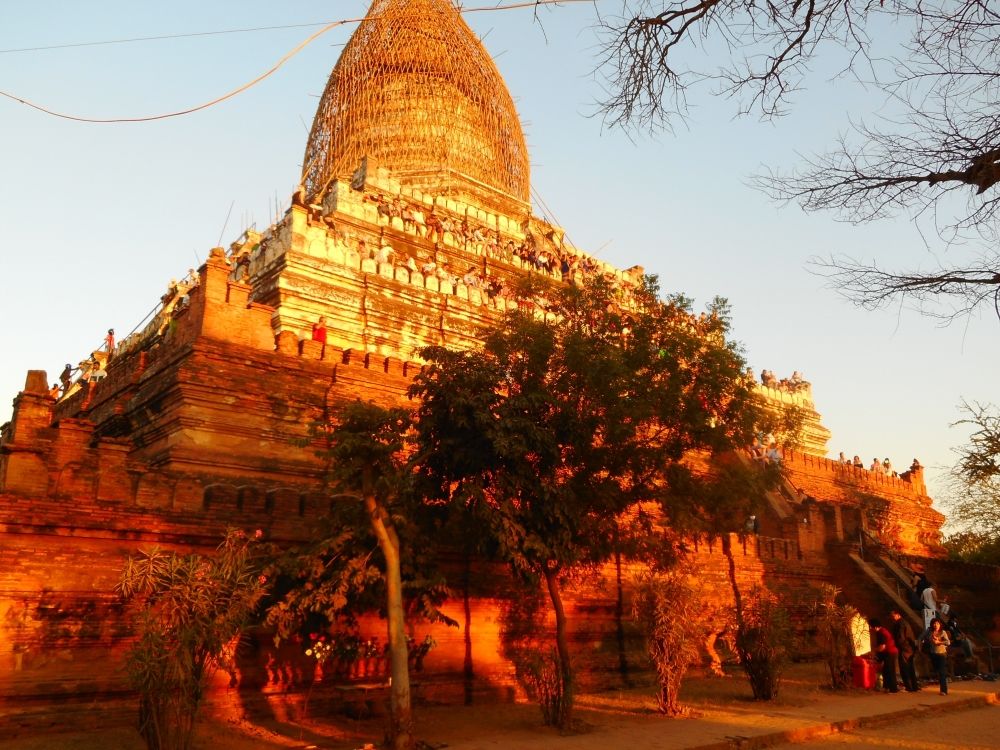 A very large, very tall temple at Bagan in Myanmar, with a stupa on its top, which seems to be enclosed in a scaffolding of some sort. The temple looks golden in the low light.