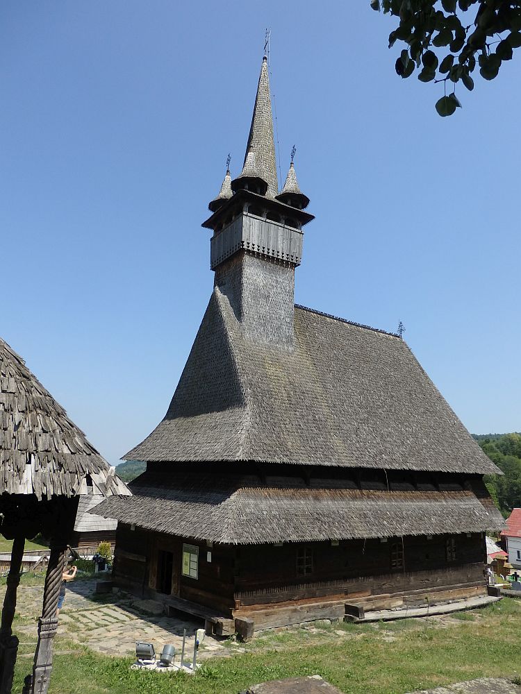One of the wooden churches of Maramures, this church has a steep double roof in neat overlapping shingles. At one end of the peak is the steeple, square at the bottom and shingled, with what looks like it might be a place to look out of at the top of the square part. Above that the steeple is round and progressively narrower. Around the base of the round part are 4 small turrets, also round and tapering and also shingled.