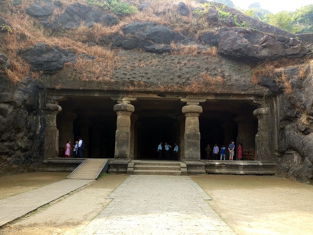 The entrance to the cave is set into the side of a hill. The cave is quite large, perhaps 2-3 stories tall, judging by the people standing right inside the entrance., and has four very large square columns holding up the wide rectangular entrance.