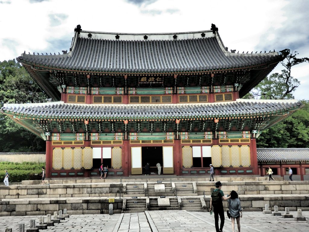 A large ceremonial hall at Changdeokgung Palace: 2 levels of curved roofs, typical of traditional Korean buildings. The walls and window frames below the lower roof and between it and the top roof are painted in bright colors. Stone steps lead up to the ground floor.