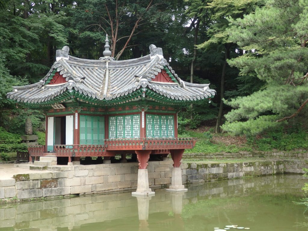 A pavilion in the garden at Changdeokgung Palace: a small room with the typical curved roof lines, green painted screens on the side. It extends out over a pond on 2 pillars.