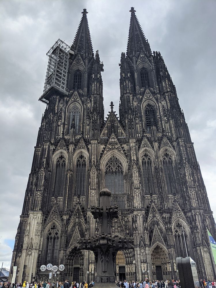 Front view of Cologne cathedral: narrow and soaring with 2 spires. Lots of large gothic windows with detailed sculptural work around them. People, very small at the bottom of the photo, lend scale to the building.
