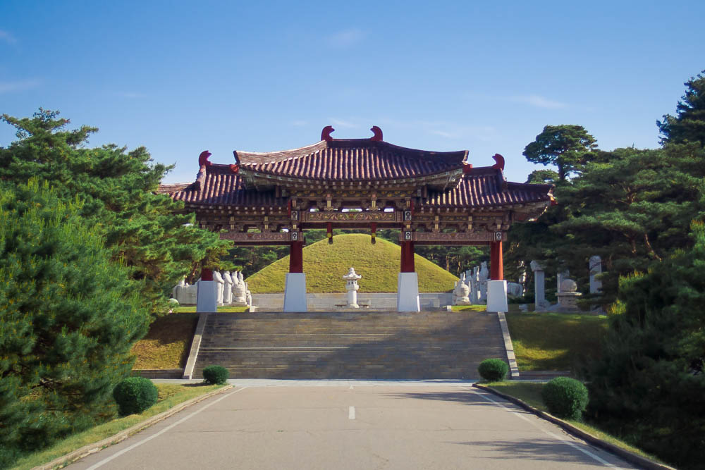An ornate gate (without doors): red roof with curved roof lines, supported by 4 red pillars, with decorative painting under the roof. Beyond that, a neat grass-covered mound.