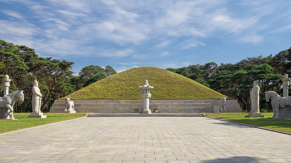A neat, grass-covered mound. Statues of various sorts in white stone line the wide path toward the mound.