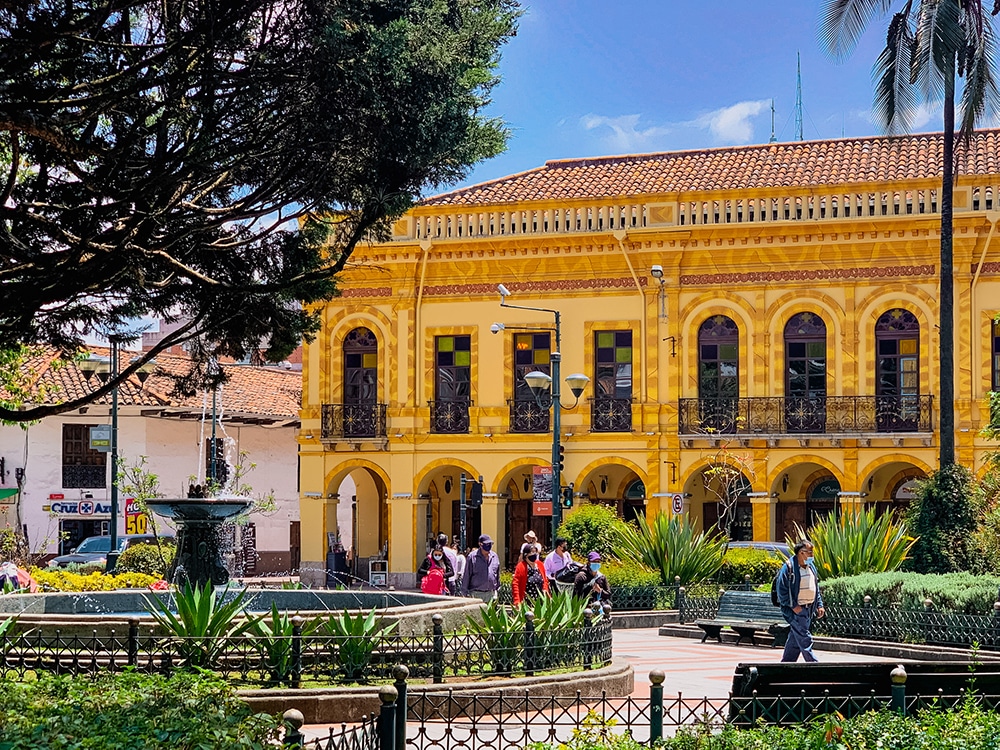 A pretty yellow building with two storeys facing on a pleasant square with greenery and a fountain.