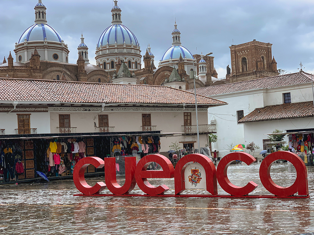 In a square where it recently rained, large letters stand that read "cuenca". Buildings on the edge of the square are just 2 storeys high, but looming above them are 3 tall domes, all blue.