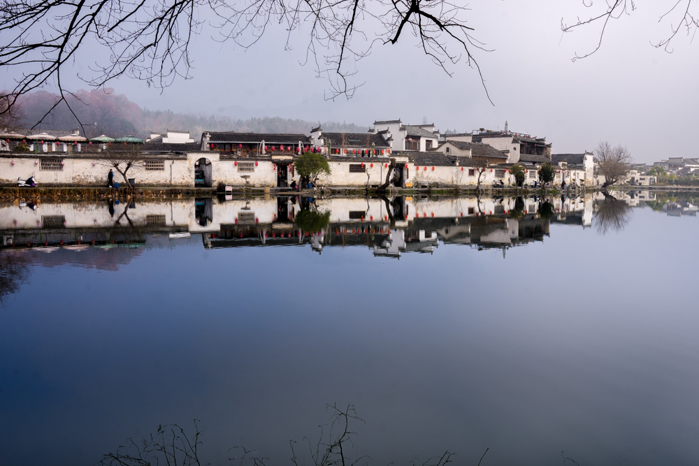 A row of low white buildings with occasional archways in the wall in front of them sit on the edge of a lake. The lake water is absolutely still, so the houses are mirrored perfectly in the water.