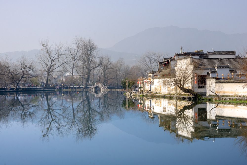 On the right, a row of low white houses on the edge of a lake. Between two of them, a tree with no leaves leans steeply out over the water. Straight ahead, just the still water, then, in the distance, an arched bridge curves over the water, and is mirrored in the water. The trees on the far shore are also mirrored in the water.