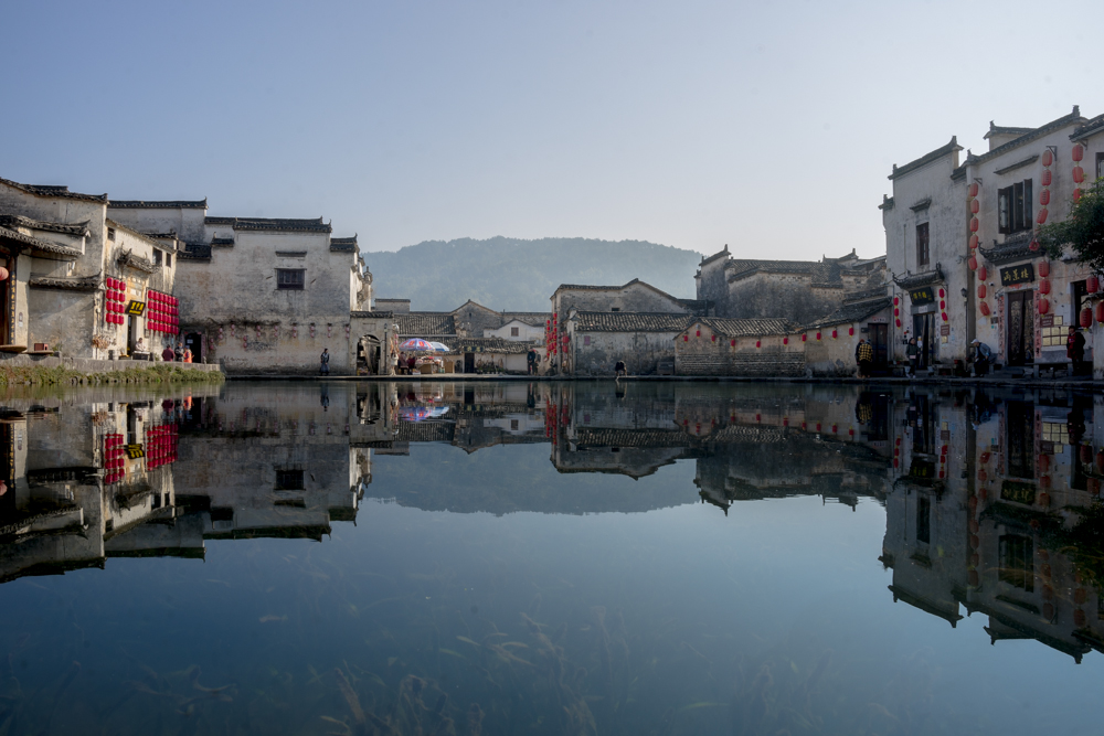 Buildings clustered along the edge of a lake. The buildings are one or two storeys tall and right up against each other. They're all white with black trim around roofs and windows, and some are adorned with bright red paper lanterns.