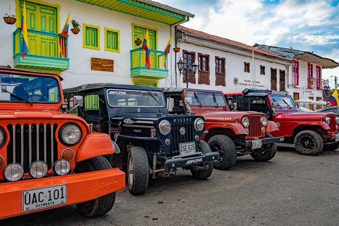 A row of parked jeeps: 3 are red, one is black. They look quite heavy-duty. Behind them are some houses or stores, in white with colorful trim.