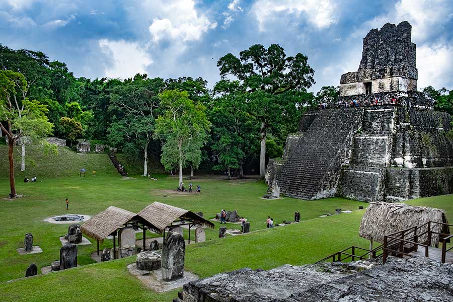 On the right, a stone step pyramid, quite steep, with a very steep stairway to its top. The top is shaped like a huge bench with a back. In the grassy area in front of the pyramid are a scattering of other smaller structures or just single large stones.