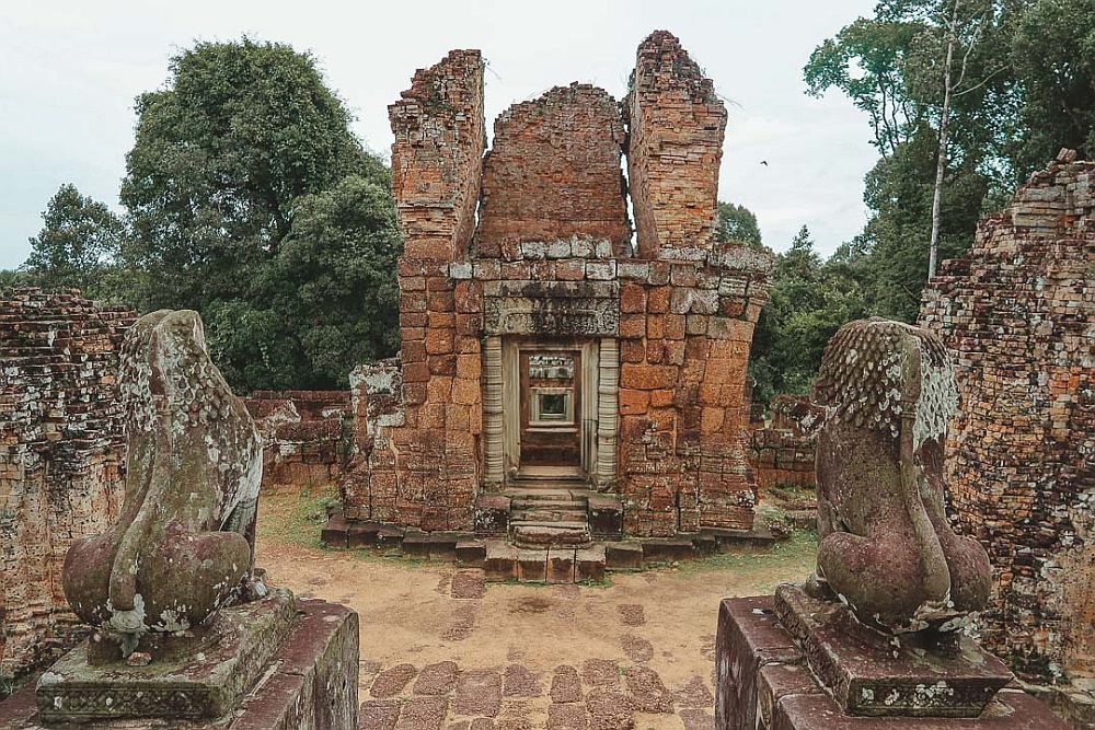 A small structure where the doorway through the middle is still intact, but the structure above it has only three partially surviving walls of brick and stone. In the foreground, two lion statues stand on either side, facing the central structure.