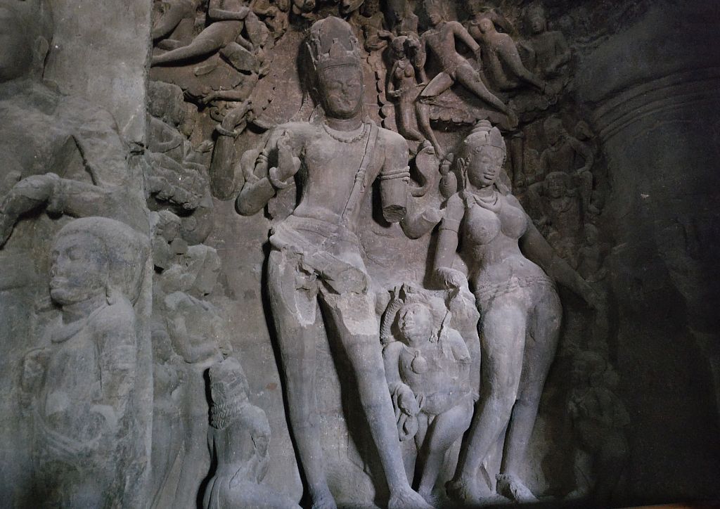 The central male Shiva figure appears to be dancing, with one armed raised in front, the other has broken off. A smaller figure next to it, female, seems to also be dancing. A multitude of both male and female figures, much smaller, surround the two main ones.