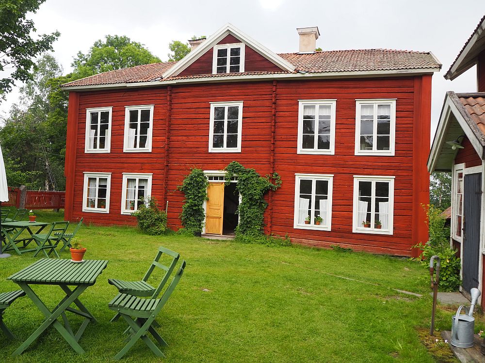 A red wooden farmhouse, symmetrical, with white trim and a yellow doorway on the entrance right in the middle. A small gable above the doorway has a 3r'd story window but most of the house is 2 stories. Green lawn in front.