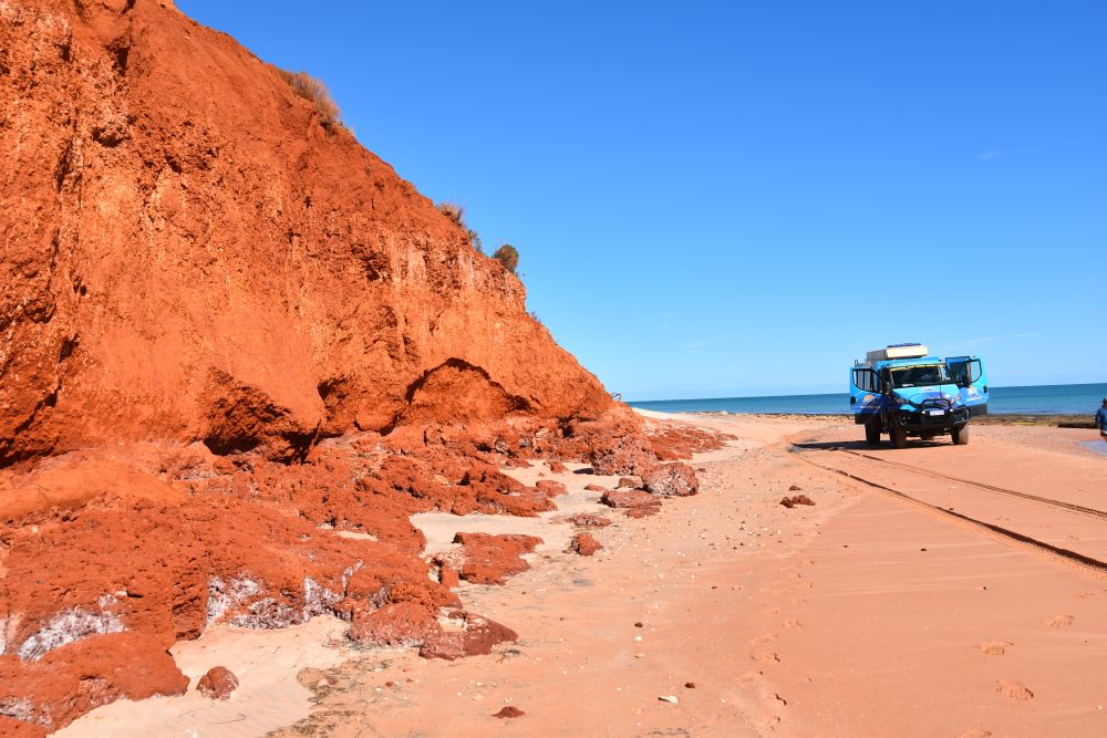 A shoreline of reddish sand, looking down the beach with water on the right and a red cliff on the left. A camper van is parked on the sand.