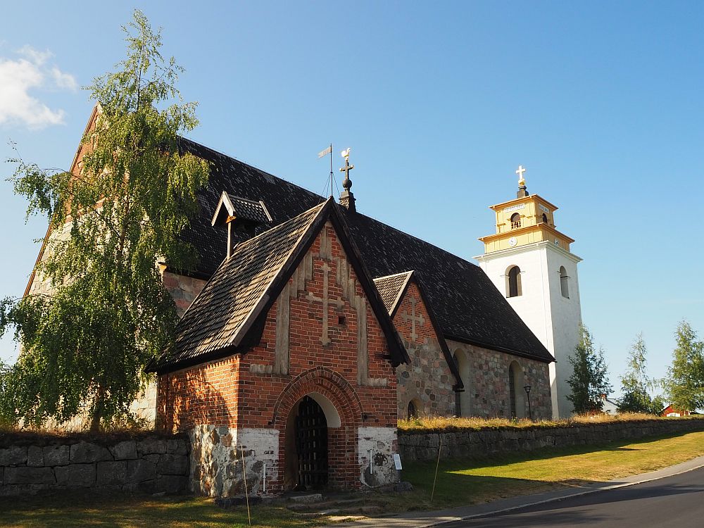 The church is partly grey stone and partly red brick. The entrance gate is at the side, with an arched entranceway. At the far end is a square white church tower.