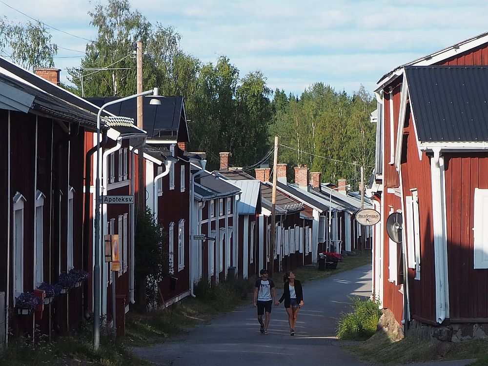 looking along a road between two rows of small red houses with white trim. The road curves out of sight. A couple, holding hands, walks on the road.