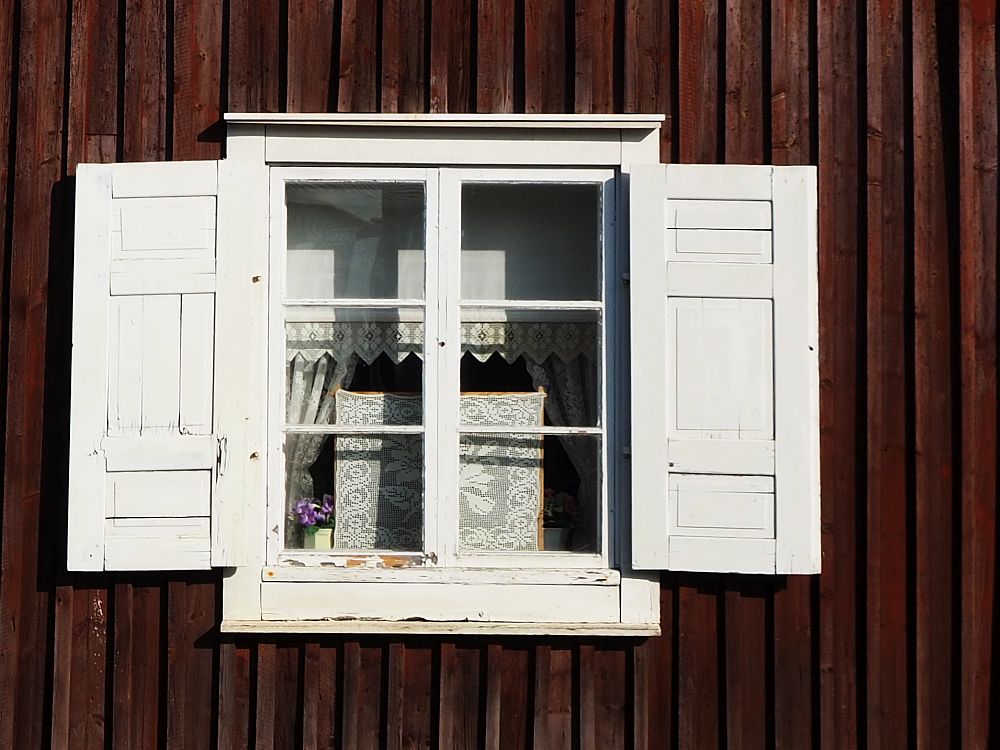 Set in a dark red-painted wall, the window has 6 panes and the trim around it is very simple, painted white. Shutters, also white, on either side.