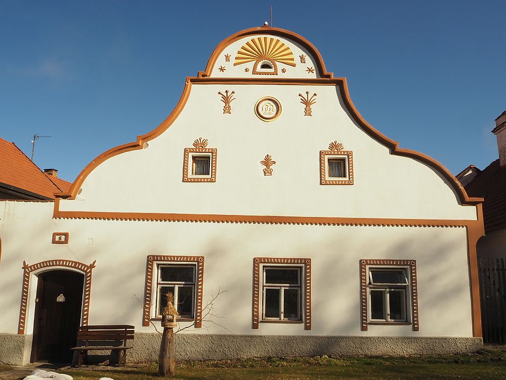 One of the Holasovice houses: Its gable is rounded on the side, and its top is a half circle. Inside the half-circle top is a decorative element with orange and yellow rays extending out from the tiny window. There are three sculptural elements on the story below that looking like, perhaps, sheaves of wheat. The ground floor is plain except for a simple painted trip around the three windows and the door.