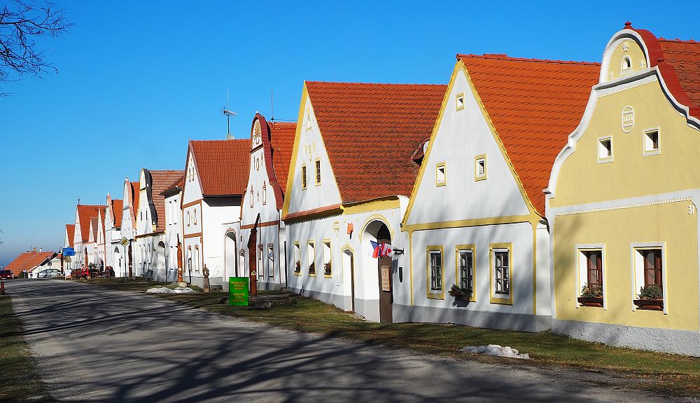 Looking down a row of houses in Holasovice. Most are white, and have trim in pastels. The gables all face the street in front of them, and vary in shape, some pointed, some rounded at the top. They are all 2 stories tall, but with an additional small window in the top of the roof.