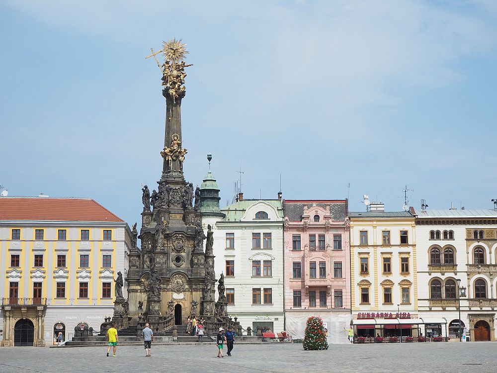 The Holy Trinity Column in Olomouc on the plaza in front of a row of elegant buildings. It is wide at the bottom, gratually narrowing until about halfway up. Above that is an almost straight column with a cluster of gold statuary at the top.