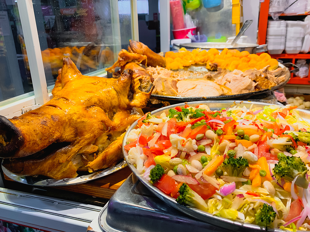 Food on display. On the left, a pig's whole head, with some sort of coating that it was cooked in, but still recognizably a head. On the right, a vegetable platter with tomatoes, broccoli, onions and beans. Behind, a tray of meat.