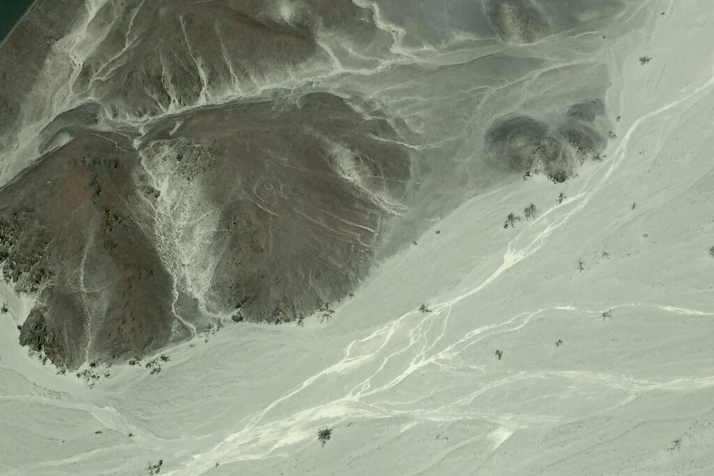 Seen from above, a low mountain, with visible stream beds, but the mountain and the flatter ground near it are all in shades of gray. A few trees here and there, from this high up just dots. Very vaguely on the side of the mountain, a simple human stick figure can be seen.