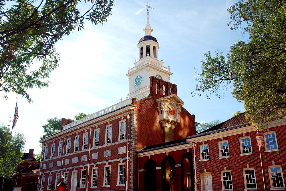 A full view of Independence Hall: red brick, two stories, with a white tower at one end of the roof. The tower's square base has a clock on both visible sides. A more ornate clock is on the end wall of the building.