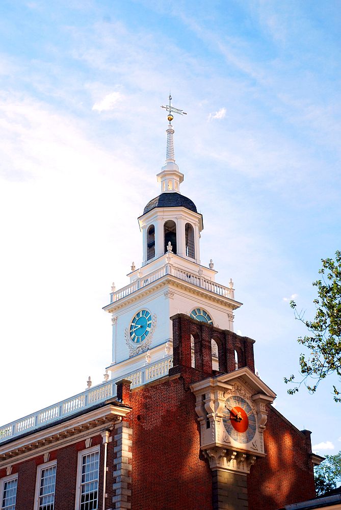 A view of the steeple on Independence Hall: the building is brick but the steeple is white, square at the base, round above, with clocks on the square sides.