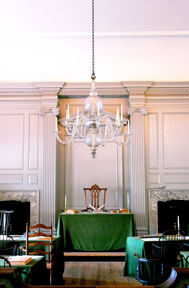 Inside the hall, looking toward a simple desk with a chair behind it and a green cloth over it. On either side of the aisle, rows of chairs, also behind green-covered tables, all chairs facing the central table. 