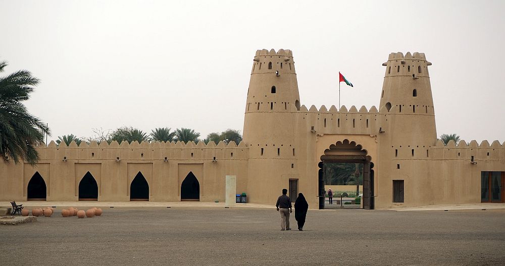 A terra cotta color entranceway with a tower at each side of it. The towers are cylindrical but taper narrower at the top. Extending out on either side of the arched entryway is a wall with pointed crenellations and arched openings that do not open all the way through the wall. 