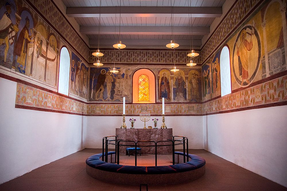 Inside the church in Jelling: the top half the walls on the 3 visible sides of the altar are painted with religious imagery and decorative borders. The altar is surrounded by a low stone circle and a railing inside of that.