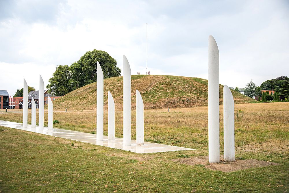 A modern sculpture in the foreground: white cylindrical poles with points pointing upwards, two different heights. Behind that, a mound, covered in grass. A few people, looking very small, stand on top of the mound.