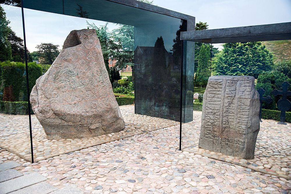 Two of the Jelling runic stones, both large rocks set into a rock-paved floor. Each has its own glass-sided case. The left-hand one has swirly images, the right has runes.