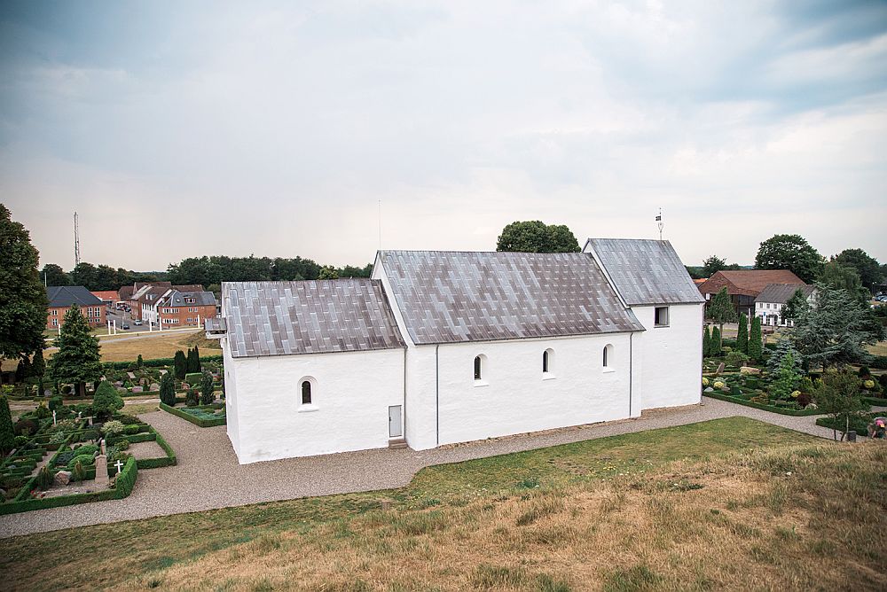 The church at Jelling is white and is made of 3 rectangular parts set end to end. The one on the right is a bit squarer than the other two and a bit taller and has a small cross on its peaked roof. There are few windows and they are small: one on the left-hand part, 3 on the middle and one on the right-hand section.