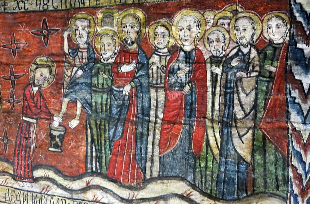 An image of Jesus washing the feet of the apostles. The Apostles are grouped on the right, int robes of blue and capes of red or white. They have halos around their heads. Jesus is on the left, washing the feet of a man who is presumably sitting, but the painting is rather unsophisticated so the man just looks smaller than the others, who are standing.