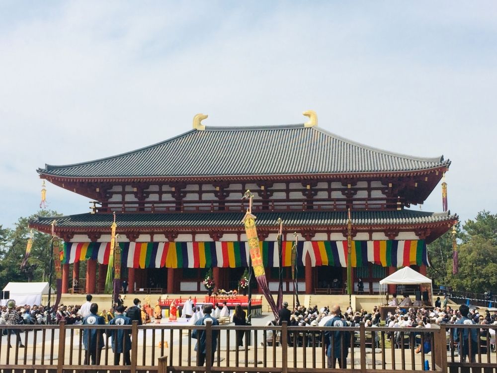 A large building with the typical curved roof, both on top of the building and around its middle. Bright banners hang under the lower eaves and wave in the wind. Some kind of ceremony is going on in the open area in front of the building.