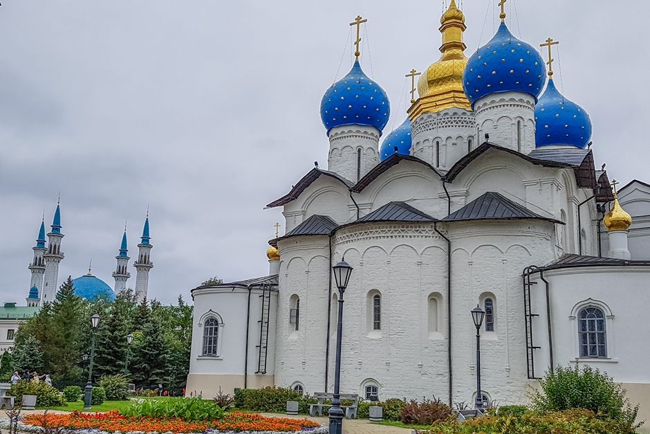 A Russian Orthodox church, with a rather simple white facade, but topped with a cluster of onion domes painted blue with small stars and a central steeple painted in gold. A mosque is visible in the background: just its central blue dome and 4 corner minarets.