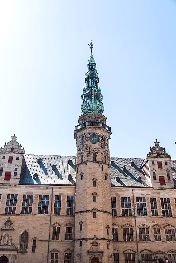 A circular tower extends outside of a flat wall of Kronborg tower. All of it ilight red brick or stone, both the wall and the tower itself, except that the top of the spire is a series of levels in green copper coming to a point at the top. The windows on the tower are very small, while the ones on the building - 3 stories under the roof - are tall rectangles. The roof looks like it holds two or three stories and 2 large, 2-story dormers are visible in this photo.