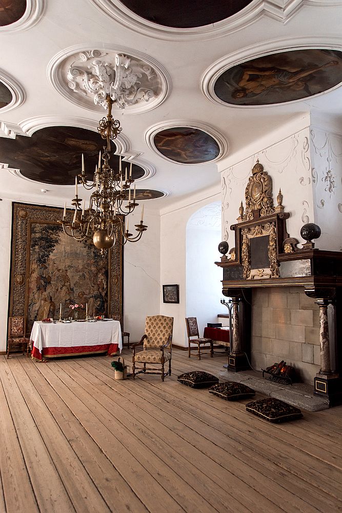 An interior in Kronborg Castle: ornate ceiling with a chandelier (with candles, not bulbs) hanging from a sculptured plaster circle and inlaid sections with dark paintings on them. On the right-hand wall, a fireplace with a baroque mantlepiece above it and plaster decorations on the white wall around it. On the far wall, a tapestry hangs and a table covered with a cloth stands in front of the tapestry.