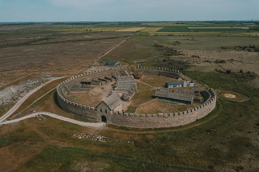 Areal view of a fortress or walled town in the middle of fields. The fortress has a brick wall around in in a circular shape, with crenellations along the top. The entrance is a building with a gateway into the wall. Inside the wall are rows of low long buildings. 