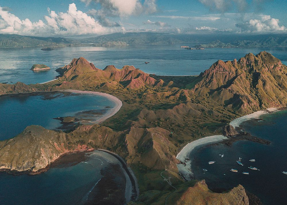 Aerial view of an island: rocky and mountainous, with round coves with very blue water. A cluster of boats are moored in one of the coves