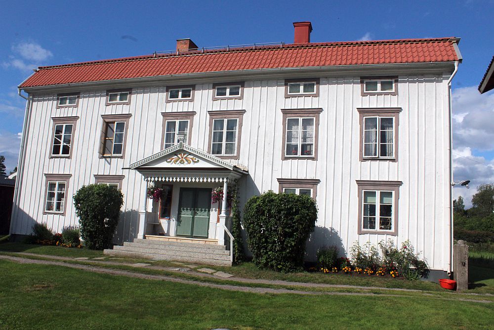 A wide white two-story farmhouse with its entrance exactly in the center, a painted decoration on the little roof above the entrance.