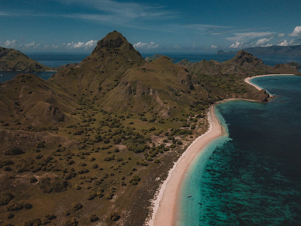 Aerial view of a stretch of mountainous land, with a beach along its edge. The water is a bright aqua color near the beach, getting darker as it gets deeper.