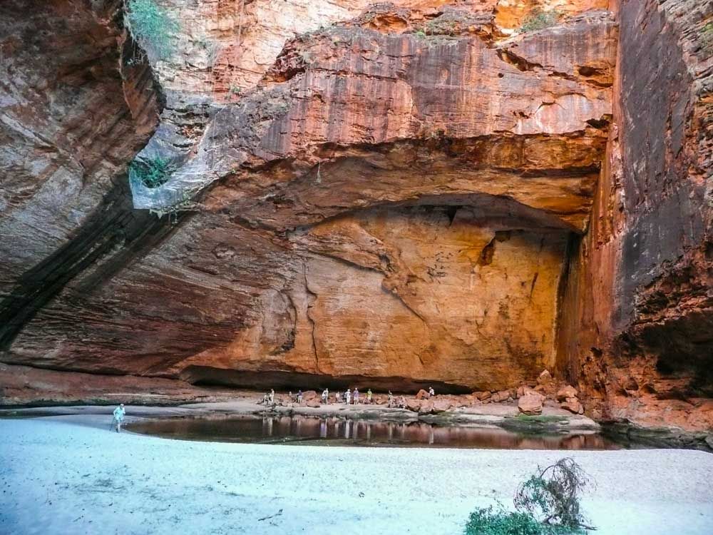 A large shallow cave in the reddish sandstone. At it's base is a small pond or puddle. Between the back of the cave and the pond people can be seen, looking very small in comparison to the huge cave.