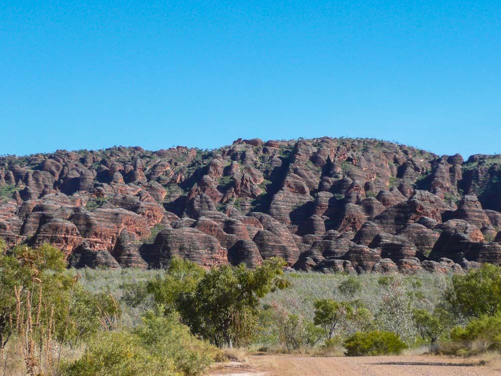 Across a grassy plain, the beehive-shaped jumble of rock formations: dark red against a very blue sky.